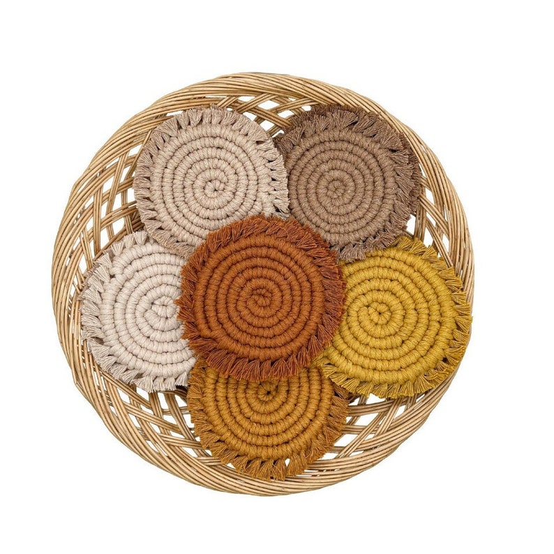 May include: Six round crocheted coasters in a wicker basket. The coasters are in shades of brown, beige, and yellow with fringe edges.