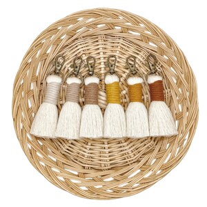 May include: Set of six white tassel keychains with different colored accents. The tassels are arranged in a circle on a woven basket.