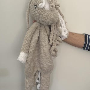 May include: A plush, beige horse-shaped baby blanket with white accents. The blanket features a horse head with floppy ears, a white blaze, and a curly white mane. The hooves and the tips of the arms are white. The blanket is held up.