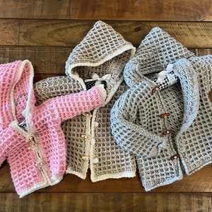 Peut inclure: Trois pulls à capuche en crochet de couleur rose, beige et gris avec des bordures blanches. Chaque pull présente un motif en nid d'abeille et des fermetures à bouton en bois.