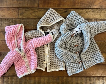 May include: Three crocheted hooded sweaters in pink, beige, and gray with white trim. Each sweater has a waffle stitch pattern and wooden toggle closures.
