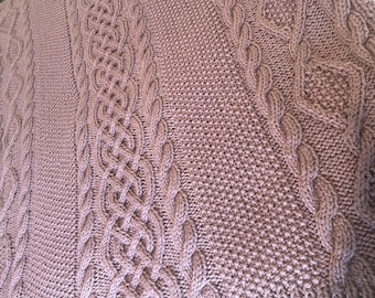 May include: Close-up of a knitted blanket in a dusty rose colour. The blanket features intricate cable knit patterns and textured stitches. The design includes vertical cable rows and a diamond-shaped cable pattern.