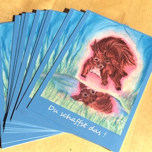 Könnte beinhalten: Ein Stapel blauer Karten mit einer Aquarellillustration von zwei Eichhörnchen und ihrer Spiegelung. Die Eichhörnchen sind braun mit rosa und blauen Akzenten. Der Text "Du schaffst das!" befindet sich unten.