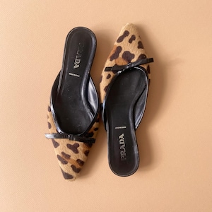 May include: A pair of Prada mules with a leopard print design. The shoes have black leather soles and small black bows. The words "PRADA" and "MADE IN ITALY" are visible on the insoles.