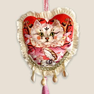 Puede incluir: Un adorno decorativo en forma de corazón con un diseño de gato, predominantemente rosa y rojo. El gato está sobre un fondo rojo, enmarcado por encaje blanco. Incluye una borla rosa, conchas marinas y detalles dorados.