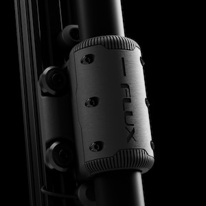 Puede incluir: Primer plano de un componente cilíndrico de metal cepillado con la palabra "FLUX" impresa en un lado. El componente está fijado a un poste negro con tornillos y una parte superior e inferior acanaladas. El fondo es negro.