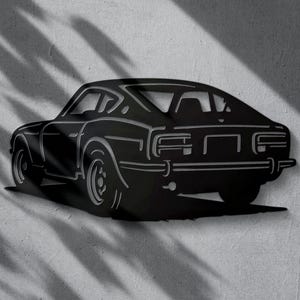 Può includere: Arte da parete in metallo nero raffigurante un'auto sportiva classica. La silhouette dell'auto è dettagliata con contorni, che mostrano le ruote, i finestrini e i dettagli posteriori. L'opera d'arte è montata su una parete grigia strutturata, con ombre che aggiungono profondità.