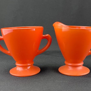 May include: A pair of vintage orange glass serving pieces. One is a creamer with a spout, and the other is a cup with two handles. Both pieces have a pedestal base and are made of a glossy, solid orange glass.