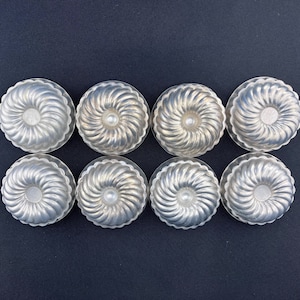 May include: Eight silver-colored metal cake molds with a swirl design. The molds are arranged in two rows of four.