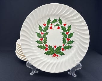 May include: White ceramic plates with a spiral edge design. Each plate features a wreath of green holly leaves and red berries. A stack of plates is visible in the background, suggesting a set.