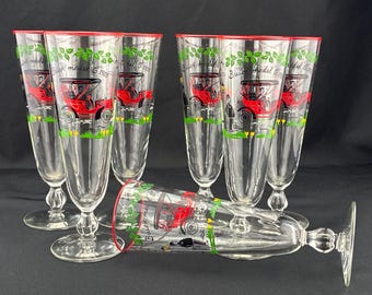 May include: A set of six clear glass pilsner glasses with red rims. Each glass features a graphic of a red 1905 Buick Model G automobile, green leaves, and the text Buick Model G 1905. The glasses have a stemmed base with a decorative knob.