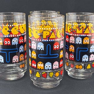 May include: Set of four clear glass tumblers featuring a colorful Pac-Man design. The glasses have a black band with Pac-Man characters and the word "PAC" in yellow. The design includes ghosts and fruit.