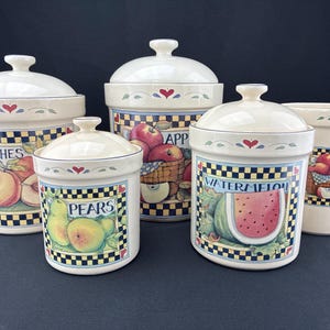 May include: A set of five ceramic canisters with lids, featuring fruit illustrations. The canisters are off-white with a black and yellow checkered pattern at the base. Each canister has a different fruit label: Peaches, Pears, Apples, and Watermelon.