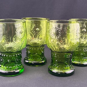 Puede incluir: Cuatro copas de vidrio verde con un dise&ntilde;o floral. Las copas tienen forma de cuenco redondo con un detallado patr&oacute;n de flores en relieve. Los tallos son de un verde m&aacute;s oscuro con un dise&ntilde;o de cuentas. Las copas est&aacute;n sobre un fondo oscuro.