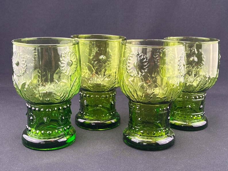 Puede incluir: Cuatro copas de vidrio verde con un dise&ntilde;o floral. Las copas tienen forma de cuenco redondo con un detallado patr&oacute;n de flores en relieve. Los tallos son de un verde m&aacute;s oscuro con un dise&ntilde;o de cuentas. Las copas est&aacute;n sobre un fondo oscuro.