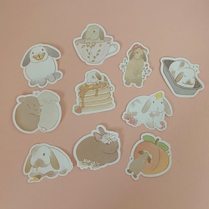 May include: A set of ten adorable bunny stickers featuring various poses and activities, including a bunny eating pancakes, a bunny wearing a mushroom hat, and a bunny sleeping. The stickers are all in a pastel color scheme and have a cute, whimsical style.