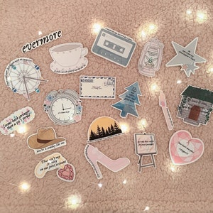 May include: A collection of stickers featuring lyrics from the album "evermore" by Taylor Swift. The stickers are on a pink fuzzy background with white fairy lights. The stickers feature various designs, including a Ferris wheel, a clock, a cowboy hat, a house, a heart, a shoe, a tree, a lantern, a cassette tape, and a star.