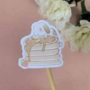 May include: A cute cartoon bunny rabbit sitting on top of a stack of pancakes with syrup and a strawberry. The pancakes are brown and the syrup is a light brown color. The bunny is white with brown ears and a brown nose.