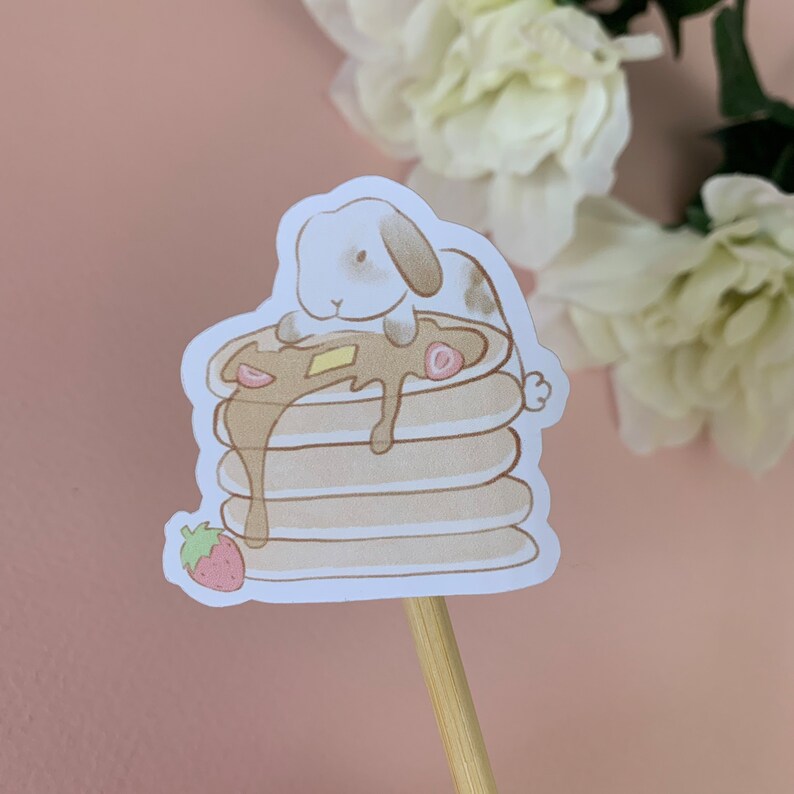 May include: A cute cartoon bunny rabbit sitting on top of a stack of pancakes with syrup and a strawberry. The pancakes are brown and the syrup is a light brown color. The bunny is white with brown ears and a brown nose.