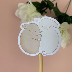 May include: A sticker featuring two brown and white rabbits cuddling. The rabbits are drawn in a simple, cartoon style. A small pink heart is drawn above the rabbits.