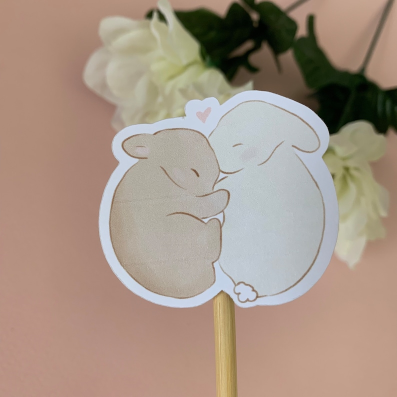 May include: A sticker featuring two brown and white rabbits cuddling. The rabbits are drawn in a simple, cartoon style. A small pink heart is drawn above the rabbits.