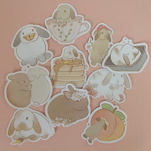 May include: A set of ten adorable bunny stickers featuring various designs, including bunnies eating pancakes, bunnies with butterflies, and bunnies holding peaches. The stickers are all in pastel colors and have a cute, whimsical style.