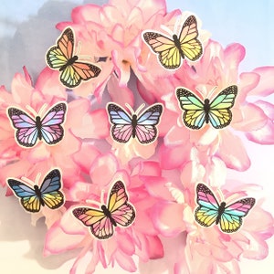 May include: A collection of colorful butterfly stickers on a pink floral background. The butterflies are in various shades of blue, yellow, orange, and green.