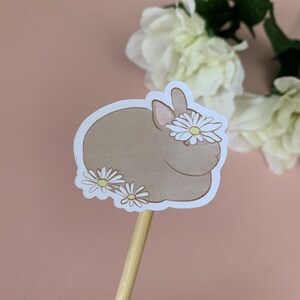 May include: A brown rabbit with white flowers on its head and around its body. The rabbit is sitting on a white background.