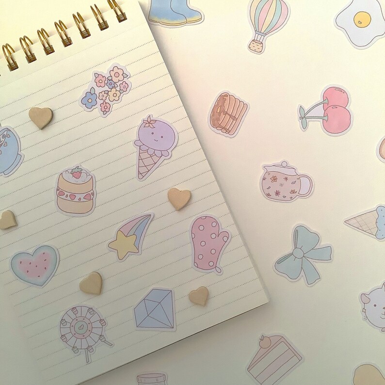 May include: A notebook with a variety of colorful stickers on it. The stickers include a hot air balloon, a star, a heart, a flower, a mitten, a cake, a waffle, a teapot, a  ice cream cone, a  mitten, a  diamond, a  ferris wheel, and a  star.