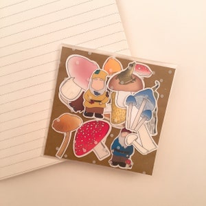 Puede incluir: Una hoja de pegatinas con setas, un gnomo, una rana y otras criaturas del bosque. Las pegatinas est&aacute;n sobre un fondo marr&oacute;n con lunares blancos.
