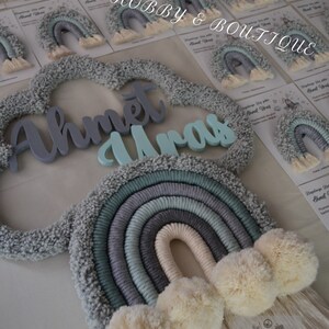 May include: A handmade rainbow wall hanging with a cloud-shaped frame and the name "Ahmet Uras" in light blue and gray lettering. The rainbow features shades of gray, blue, and cream, with fluffy pom-poms and a fringe. Small rainbow-shaped favors are in the background.