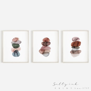 May include: Set of three abstract watercolour prints featuring stacked, overlapping shapes in shades of pink, brown, green, and blue. The prints are framed in a simple white frame.