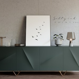 Peut inclure: Une impression minimaliste en noir et blanc d'un vol d'oiseaux en formation. L'impression est encadr&eacute;e et accroch&eacute;e &agrave; un mur au-dessus d'un meuble vert fonc&eacute; avec des pieds dor&eacute;s. Une petite plante en pot et une lampe de table sont sur le meuble.