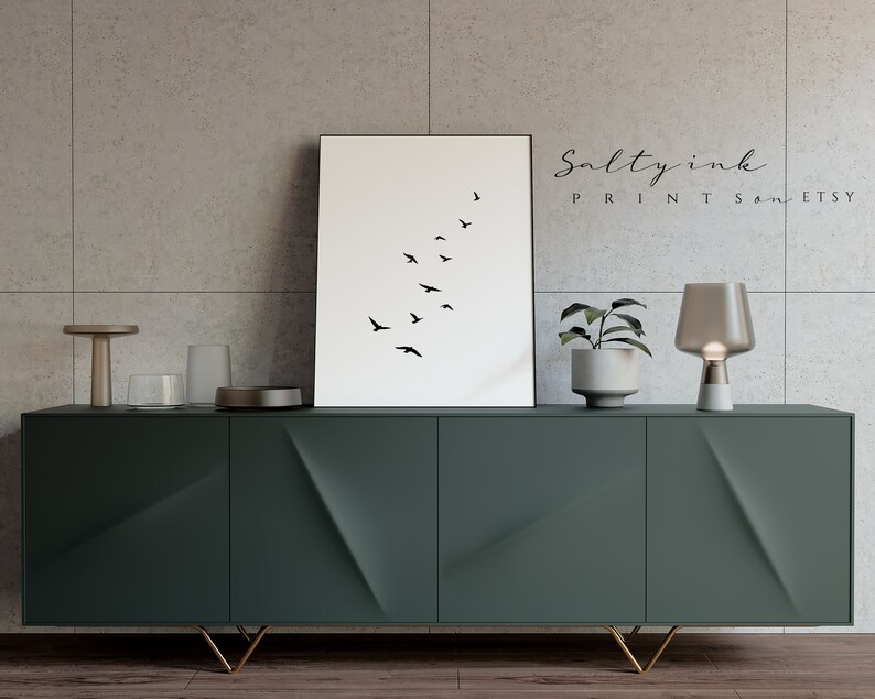 Peut inclure: Une impression minimaliste en noir et blanc d'un vol d'oiseaux en formation. L'impression est encadr&eacute;e et accroch&eacute;e &agrave; un mur au-dessus d'un meuble vert fonc&eacute; avec des pieds dor&eacute;s. Une petite plante en pot et une lampe de table sont sur le meuble.