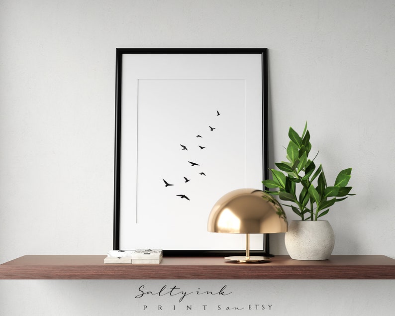 Peut inclure: Impression minimaliste en noir et blanc d'un vol d'oiseaux en formation de V. L'impression est encadr&eacute;e dans un cadre noir et est expos&eacute;e sur une &eacute;tag&egrave;re avec une lampe dor&eacute;e et une plante en pot.