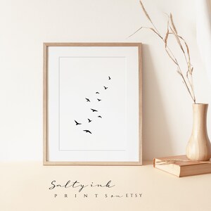 Peut inclure: Une impression minimaliste en noir et blanc d'un vol d'oiseaux en formation. L'impression est encadr&eacute;e dans un cadre en bois clair et est affich&eacute;e sur une surface beige clair.