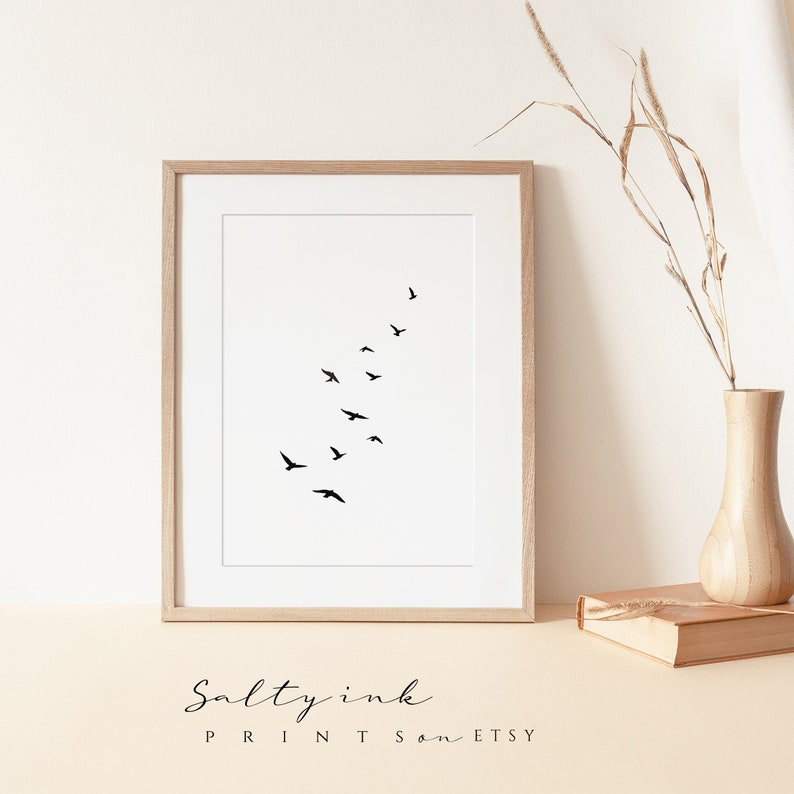 Peut inclure: Une impression minimaliste en noir et blanc d'un vol d'oiseaux en formation. L'impression est encadr&eacute;e dans un cadre en bois clair et est affich&eacute;e sur une surface beige clair.