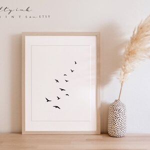 Peut inclure: Une impression minimaliste en noir et blanc d'un vol d'oiseaux en formation de V. L'impression est encadr&eacute;e dans un cadre en bois clair et est affich&eacute;e sur une surface en bois.