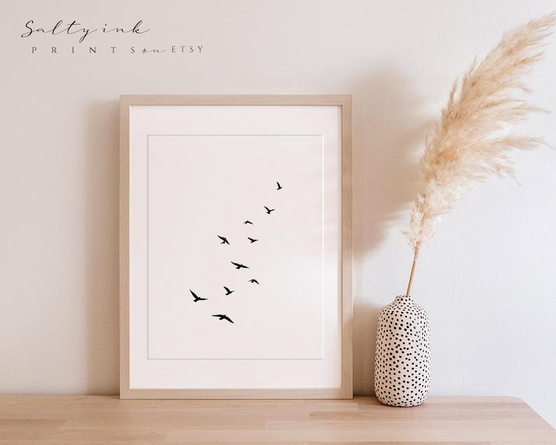Peut inclure: Une impression minimaliste en noir et blanc d'un vol d'oiseaux en formation de V. L'impression est encadr&eacute;e dans un cadre en bois clair et est affich&eacute;e sur une surface en bois.