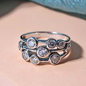 Puede incluir: Un anillo plateado con tres bandas, cada una adornada con piedras preciosas redondas y transparentes. Las piedras están engastadas en biseles individuales, creando un efecto brillante y en capas. El anillo se muestra sobre una superficie rosa claro.