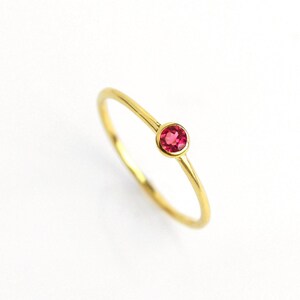 May include: A delicate gold ring featuring a small, round, bright pink gemstone. The ring has a thin band and a simple, elegant design. The gemstone is set in a bezel setting, and the ring is shown against a white background.