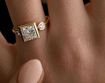 Puede incluir: Un anillo de oro con un diamante central de talla cuadrada grande, flanqueado por dos diamantes redondos más pequeños. El anillo está engastado en una fina banda de oro. El anillo se muestra en un dedo.