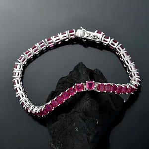 May include: Two silver bracelets with square, deep red gemstones. The bracelets are displayed on a black surface. The gemstones are set in a silver metal setting, and the bracelets have a classic tennis bracelet design.