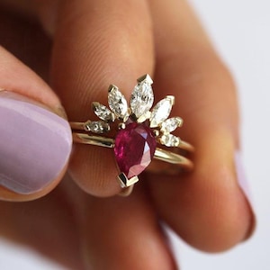 Può includere: Un anello in oro con un rubino rosso intenso a forma di pera come pietra centrale, accentuato da una corona a ventaglio di sette diamanti bianchi taglio marquise disposti in un motivo curvo e radiante sopra la gemma principale su una fascia.