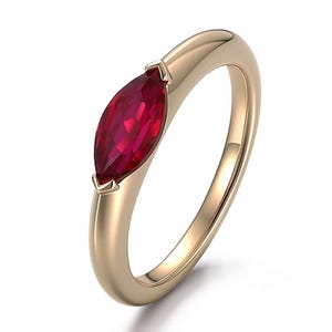 May include: A gold ring featuring a single marquise-cut ruby gemstone set horizontally. The band has a smooth, polished finish and a rounded profile, with the vibrant red stone secured by two small metal prongs at each pointed end of the gem.