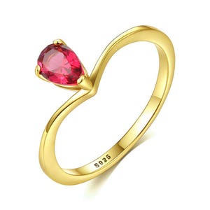 Può includere: Un anello color oro con una pietra preziosa rossa sfaccettata a forma di pera montata su griffe. La fascia presenta una curva a V che conduce alla pietra ed è incisa con il testo S925 sulla superficie interna, indicando argento sterling.