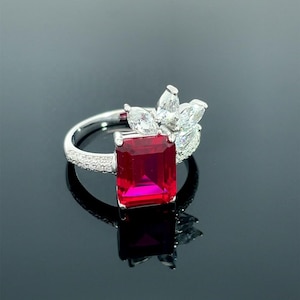 May include: A silver-toned ring featuring a large, rectangular, vibrant red centre stone. The band is accented with small, clear pavé stones, and a cluster of marquise-cut clear stones is arranged in a fan-like pattern above the red stone.