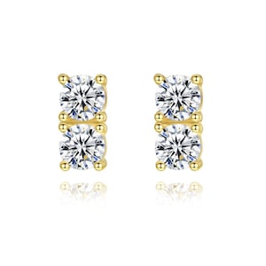 May include: A pair of gold stud earrings, each featuring two vertically aligned round brilliant-cut clear stones held in place by four small prongs. The metal setting is polished gold, and the stones exhibit clear facets that reflect light.