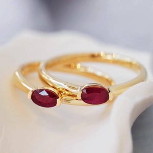 Puede incluir: Dos anillos de oro, cada uno con una piedra preciosa de rubí rojo intenso de corte ovalado engastada en una banda de metal lisa. Los anillos están superpuestos, mostrando el acabado en oro amarillo pulido y el color rojo vibrante y saturado de las piedras.