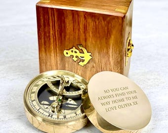 May include: A vintage-style brass compass with a hinged lid, next to a wooden box with a gold anchor emblem. The compass is open, revealing the dial. The lid of the compass has an engraved message.